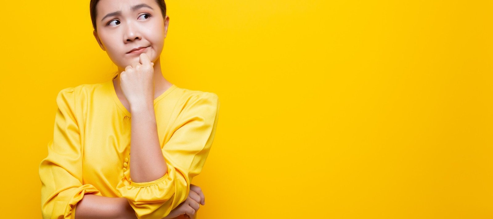 Thoughtful young woman in a yellow outfit standing against a bright yellow background, looking to the side with a curious expression wondering how to get more walk-ins.